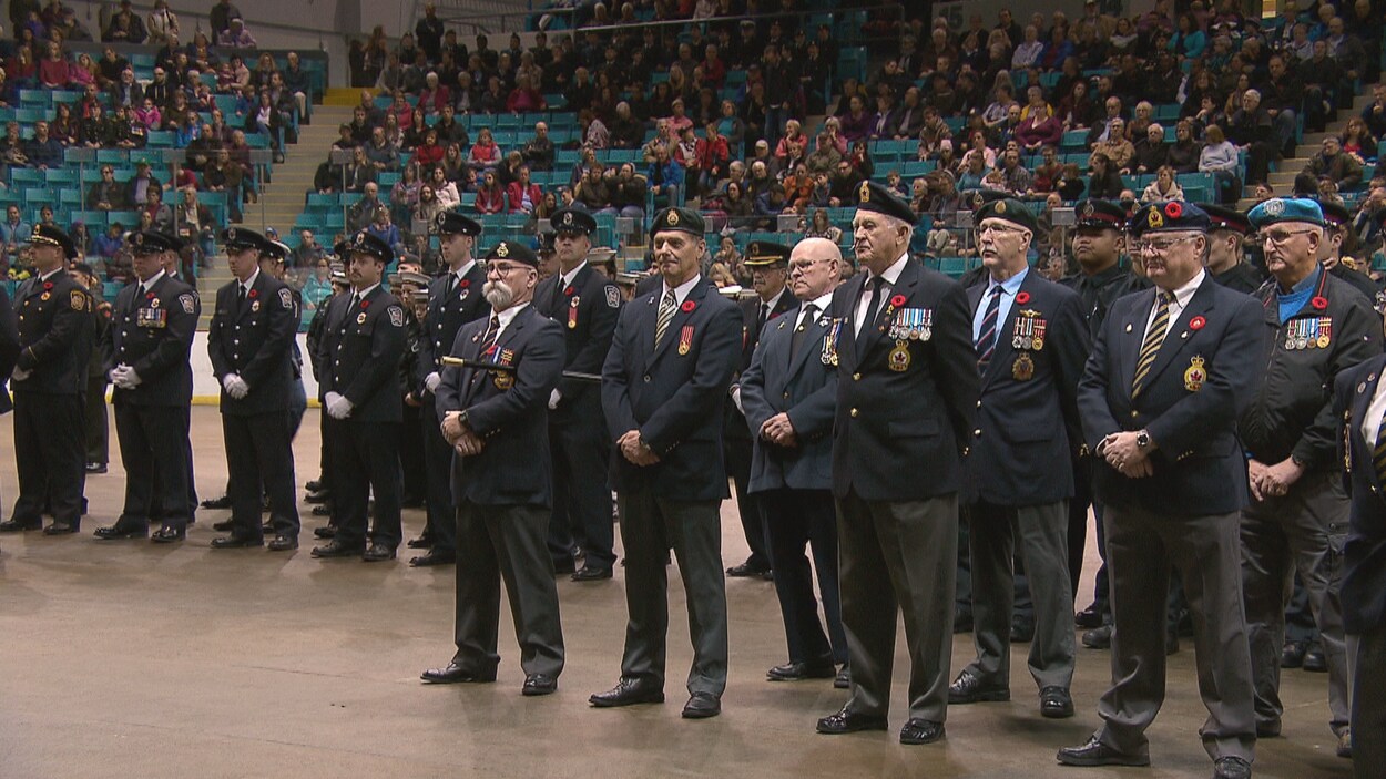 Un groupe d'anciens combattants au garde-à-vous au milieu du Colisée de Moncton. La foule occupe presque tous les gradins.