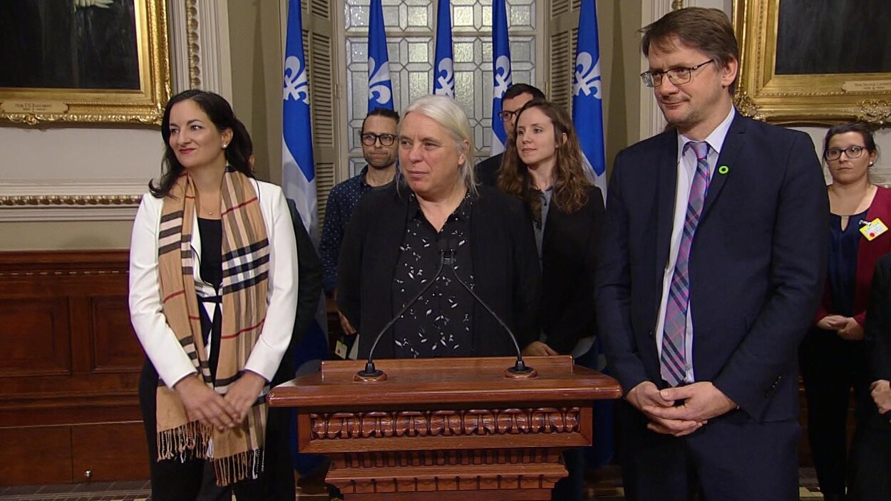 Les trois politiciens sont debout côte à côté et semble écouter quelqu'un (hors-cadre) à leur droite.