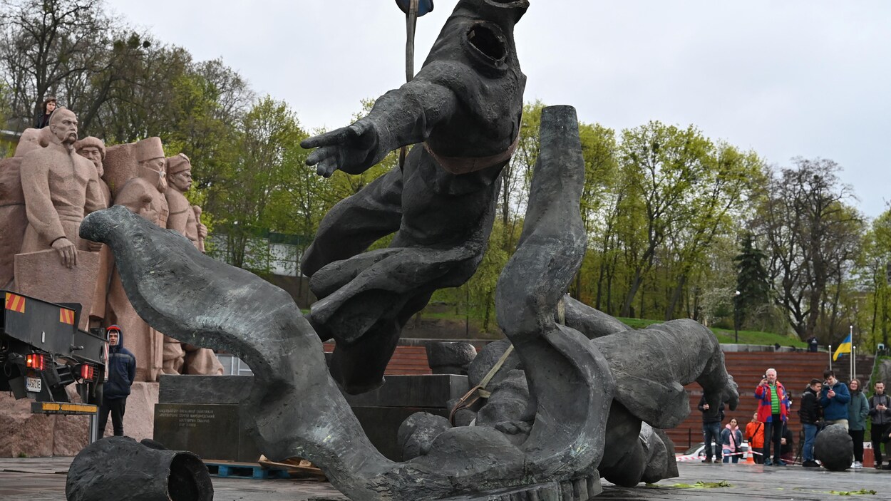 Le monument en train d'être déboulonné est suspendu à une grue sous l'oeil d'Ukrainiens, dont certains prennent des photos; les deux têtes de la statue sont tombées, mais on en voit une seule sur le sol.