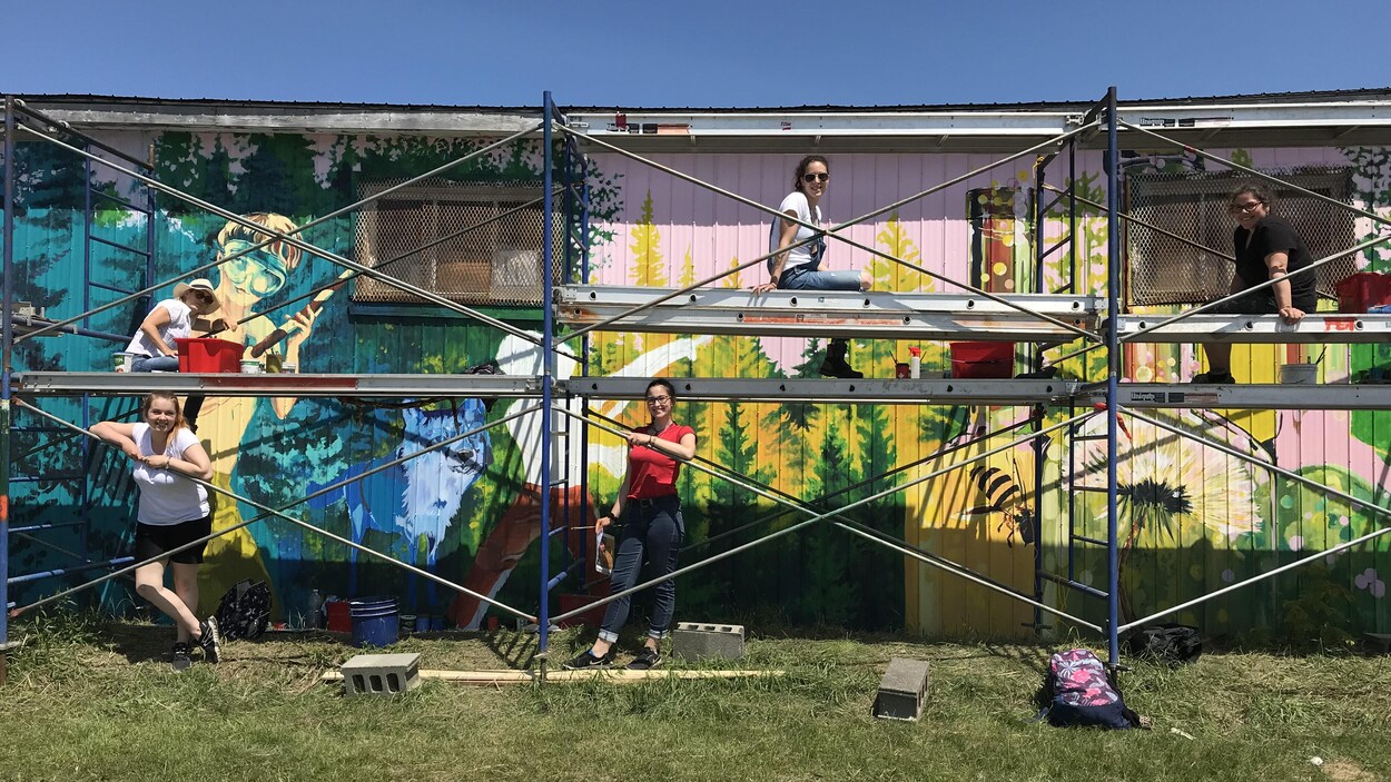 Cinq femmes sont assises dans des échafaudages devant un bâtiment où elles peignent une murale.