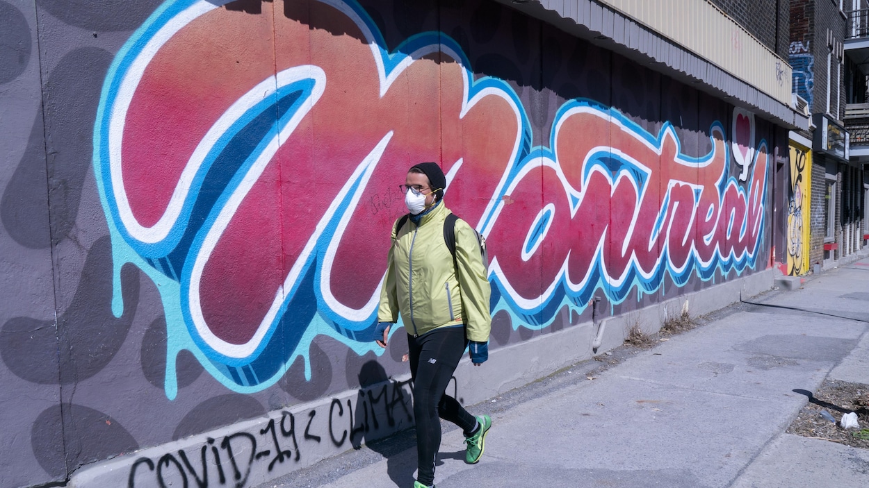 Un homme au manteau jaune portant un masque passe devant une murale sur laquelle est écrite le mot Montréal.