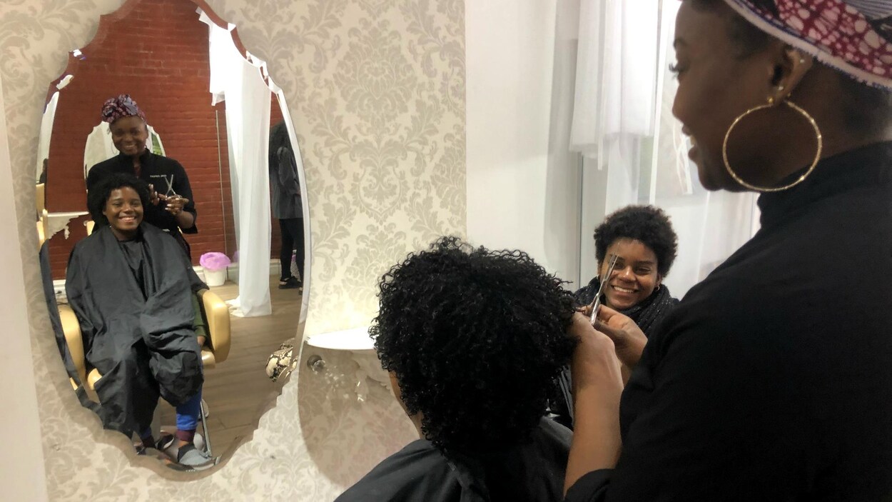 Une jeune femme noire est assise sur une chaise chez une coiffeuse et se regarde dans un miroir.