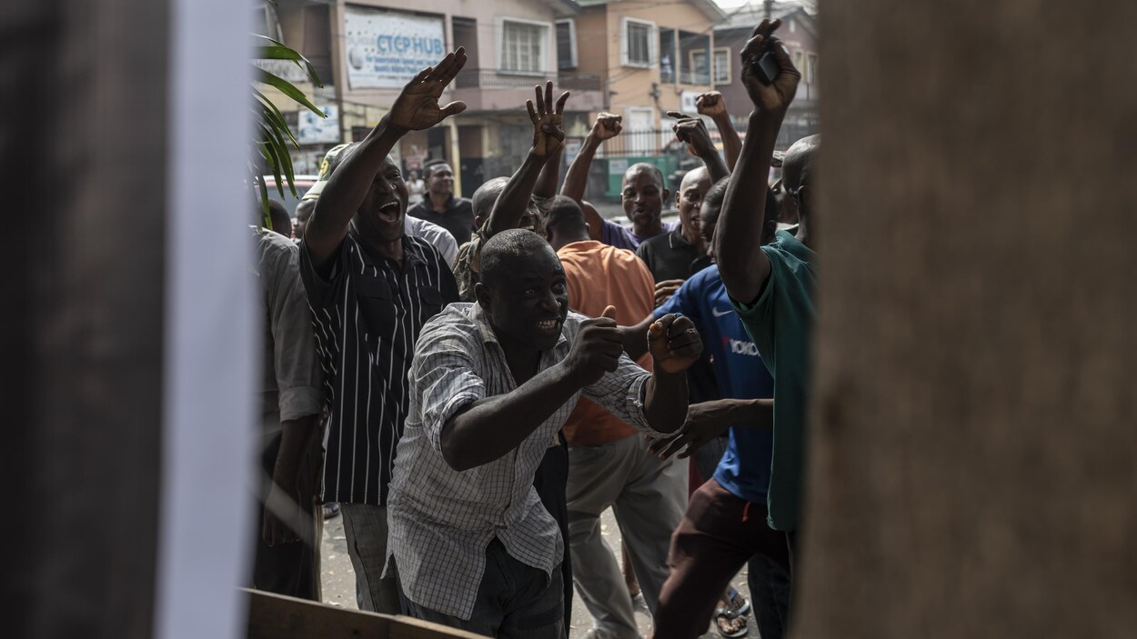 Des hommes nigérians lèvent les bras dans les airs devant un kiosque à journaux dans une rue.