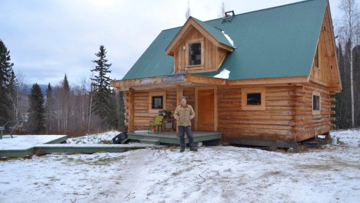 Norm Carlson est debout à l'extérieur devant sa maison en bois rond.