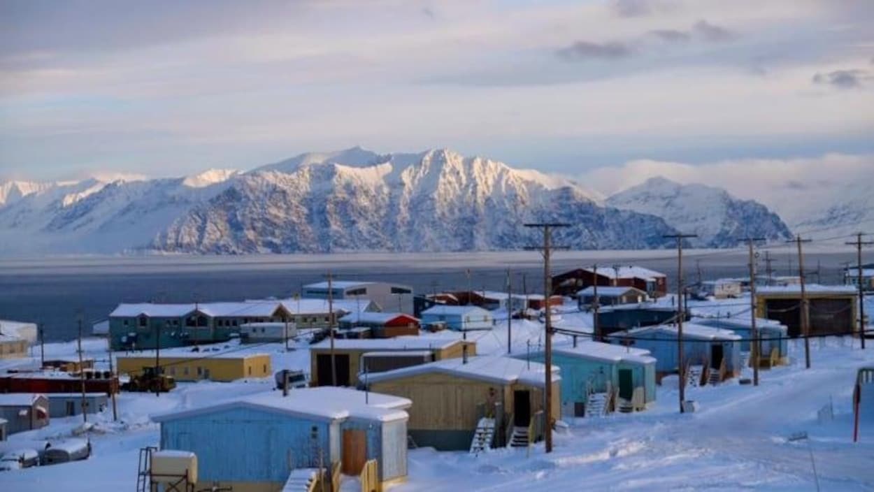 Un village enneigé devant une étendue d'eau et des montagnes.