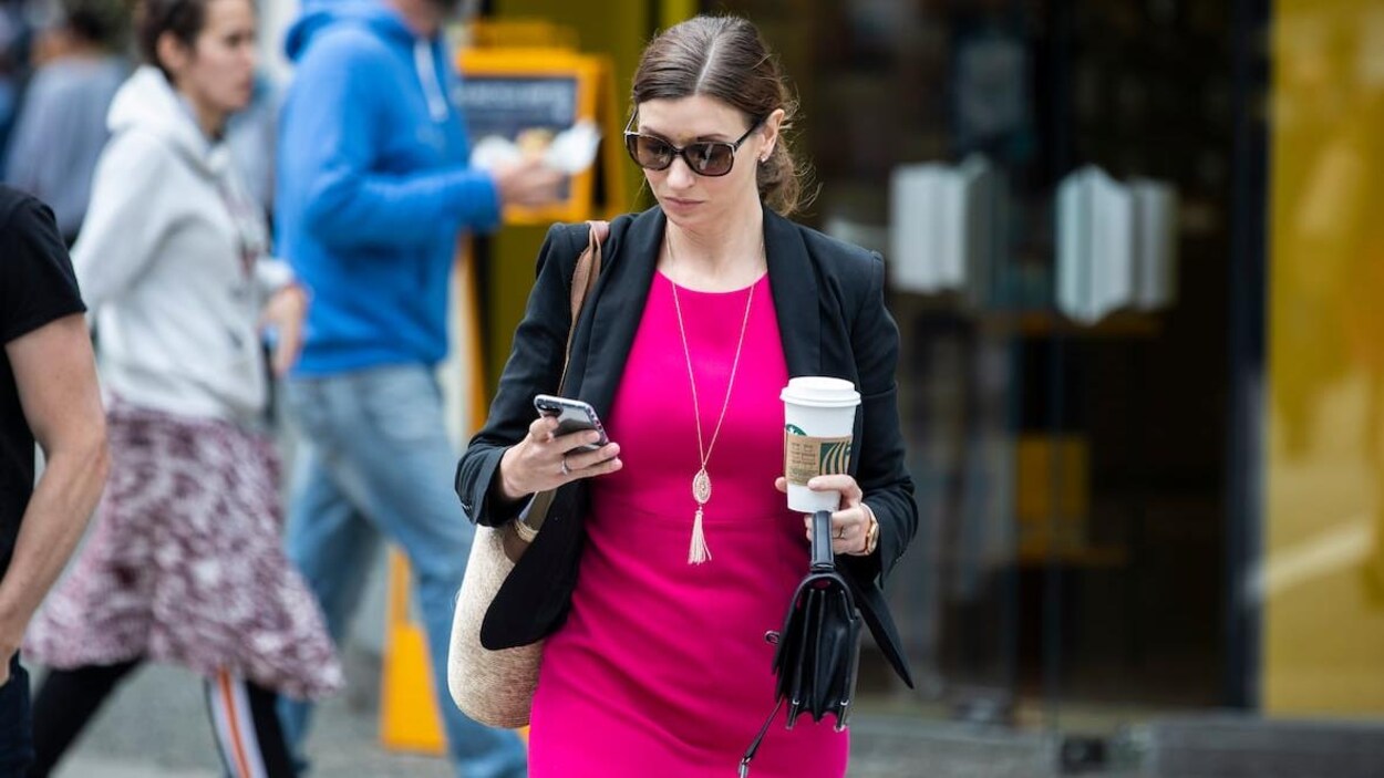Une femme portant des lunettes de soleil et une robe marche dans la rue en regardant son cellulaire alors qu'elle porte un gobelet de café dans l'autre main.