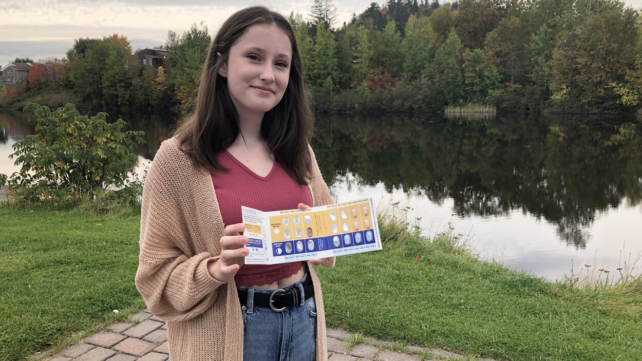 Une adolescente montre sa plaquette de médicament. Elle pose devant une rivière, en automne.