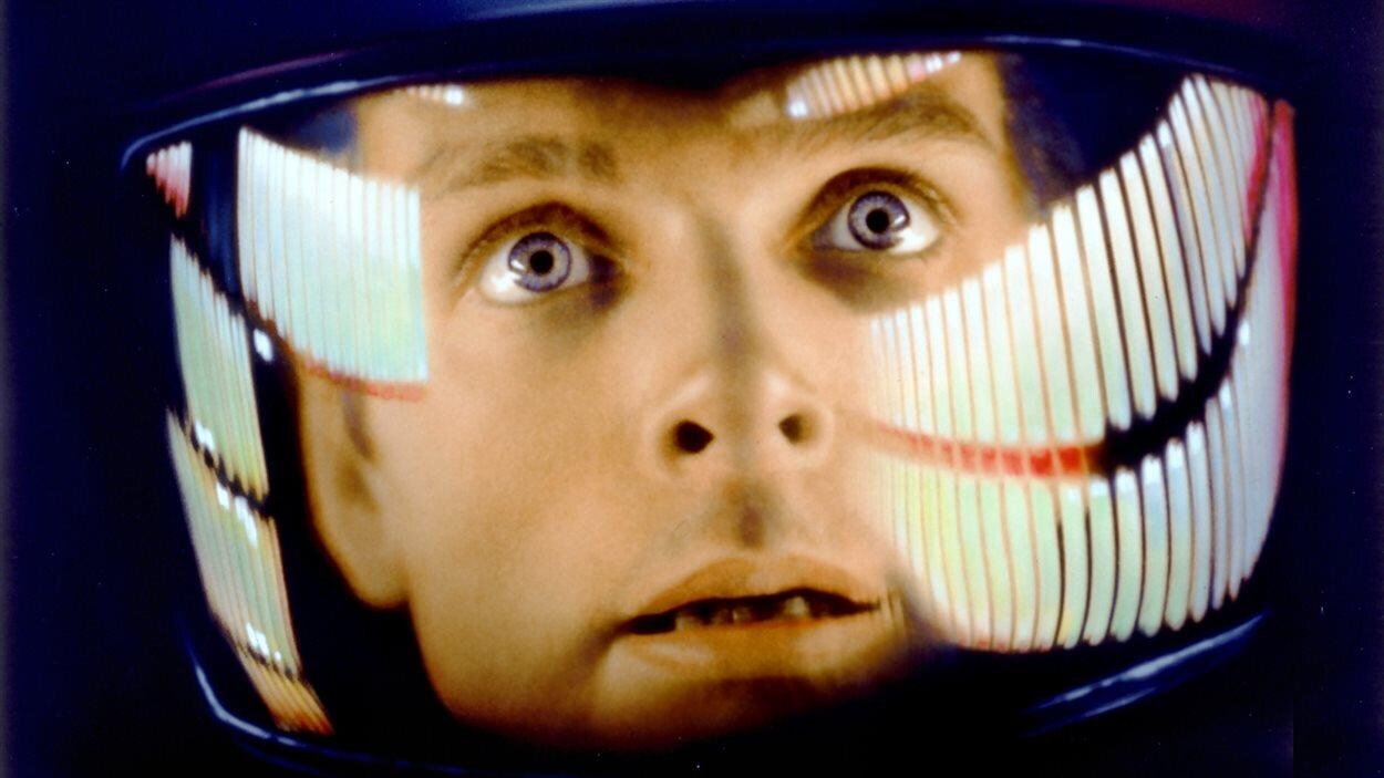 Le visage d'un homme blanc aux yeux bleus, en gros plan, regarde l'horizon. Il porte un scaphandre.