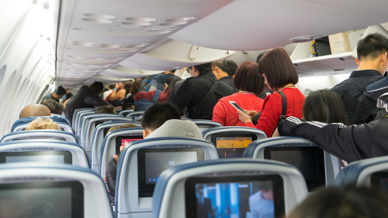 On voit, de dos, des gens entassés dans l'allée d'un avion, et d'autres assis, qui attendent pour débarquer.
