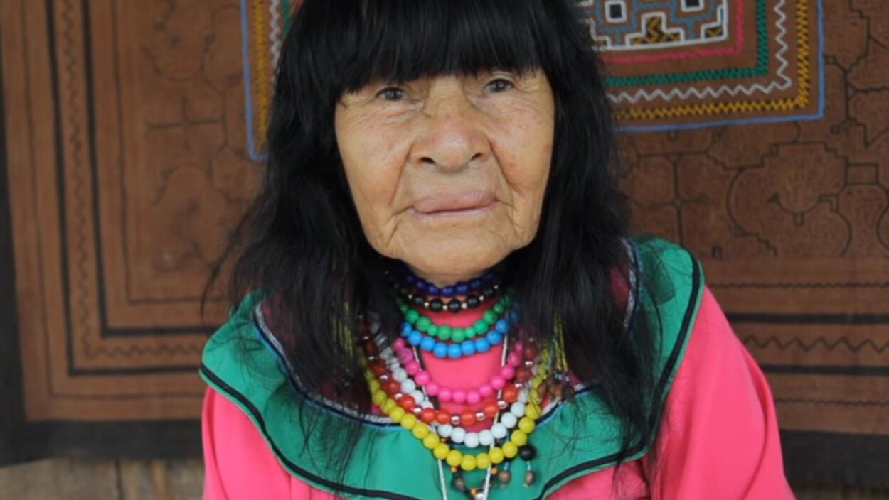 Une femme âgée habillée de couleur vive est debout devant un tissu avec des motifs autochtones.