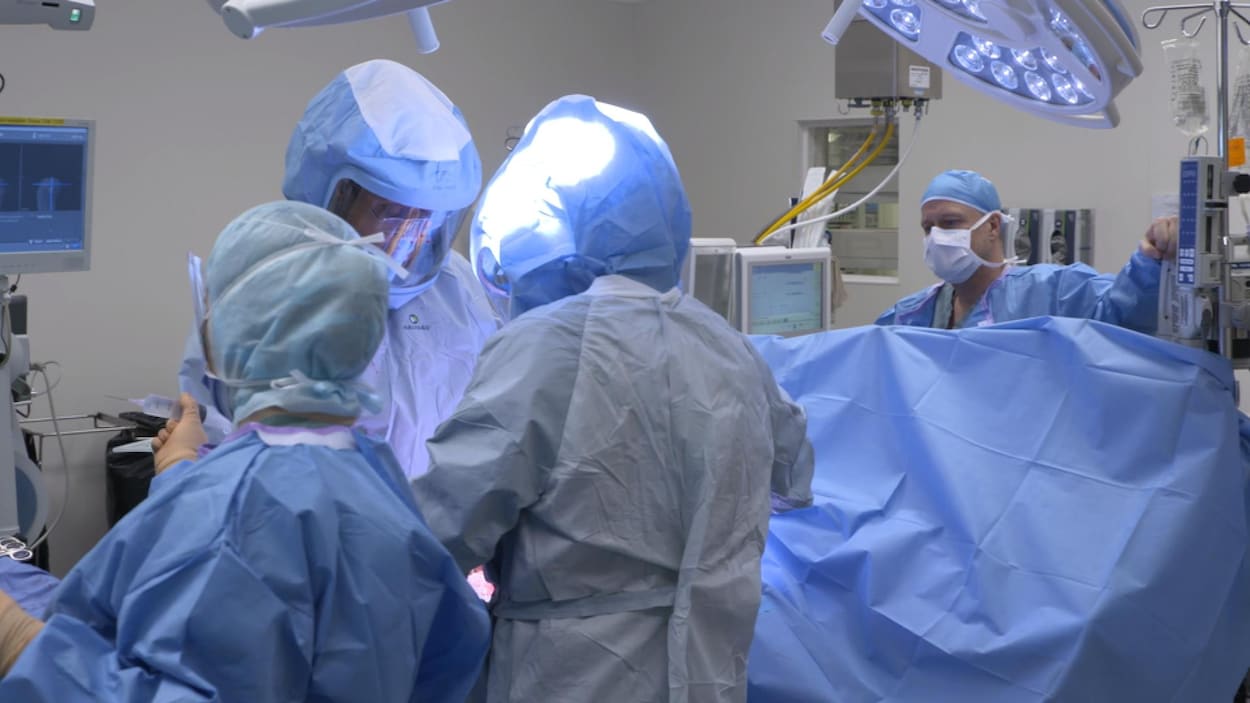 Une salle d'opération, médecin et personnel sont masqués, le patient est sous un grand drap bleu.