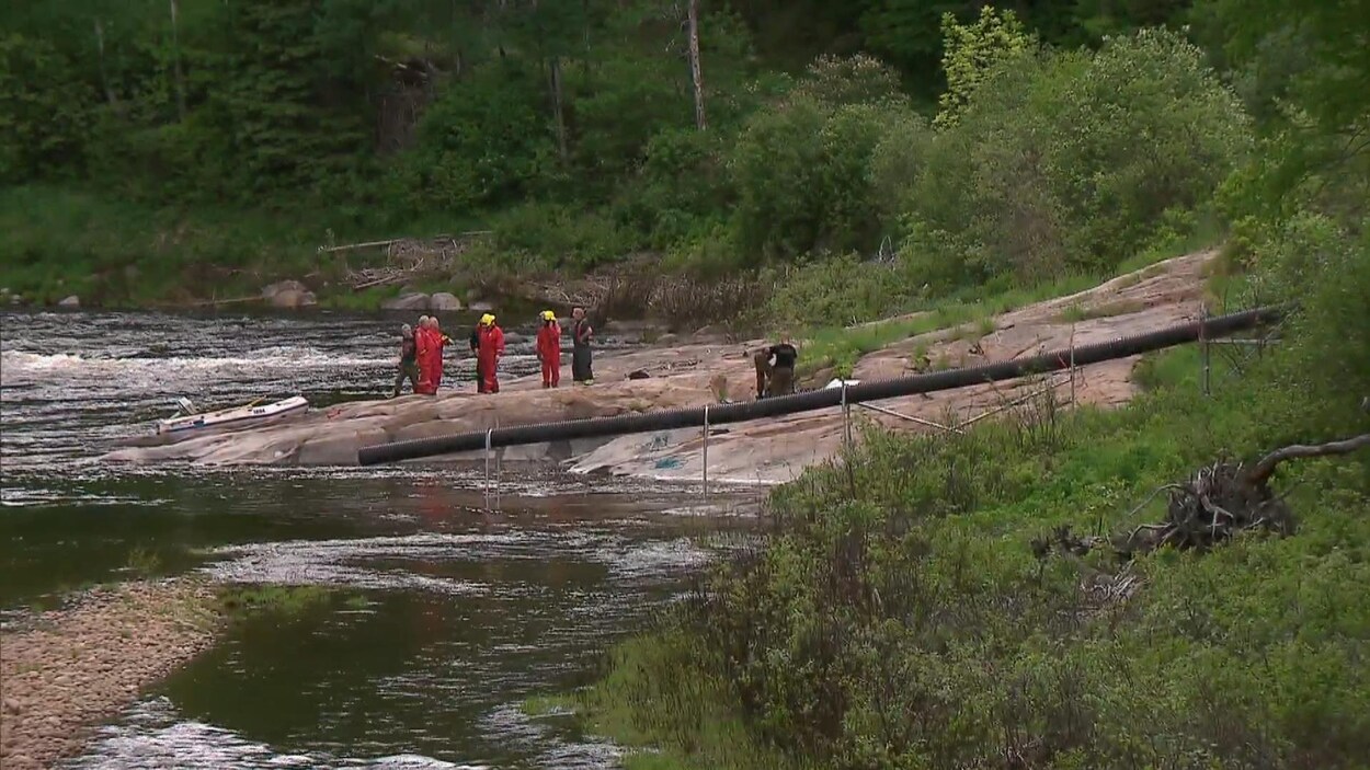 Des secouristes se tiennent sur l’une des rives de la rivière Malbaie. On aperçoit un canot pneumatique échoué sur des rochers.