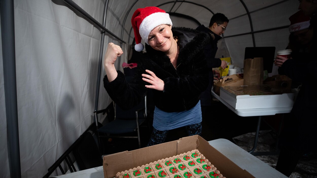 Une femme debout derrière un gâteau des Fêtes et coiffée d'un chapeau de père Noël fléchit son biceps droit en souriant.