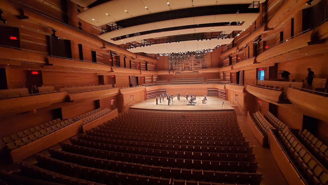 Salle de concert vide avec une dizaine de musiciens sur scène, vus de loin.