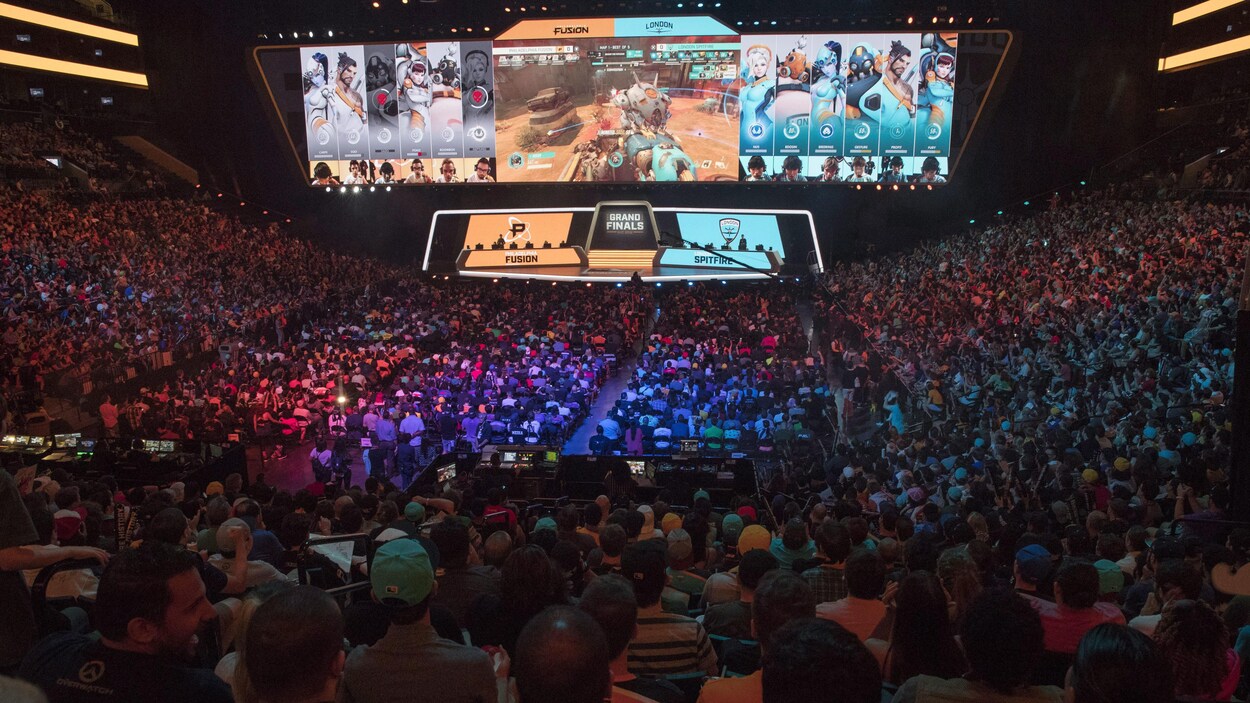 Une foule de plusieurs milliers de personnes massés dans les gradins d'un aréna regardent une scène et des écrans géants affichant une partie du jeu vidéo Overwatch.