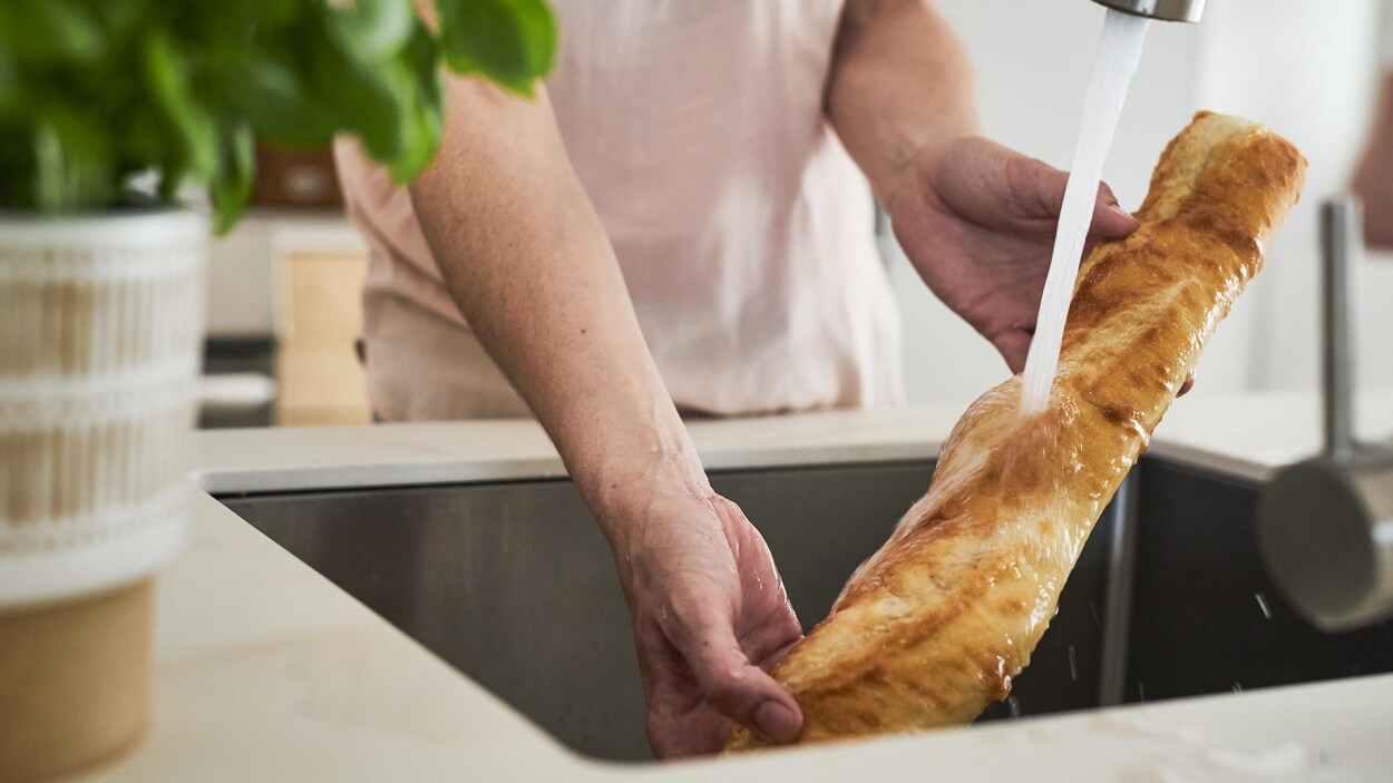 Deux mains tiennent une baguette de pain dans un évier en le passant sous un jet d'eau.
