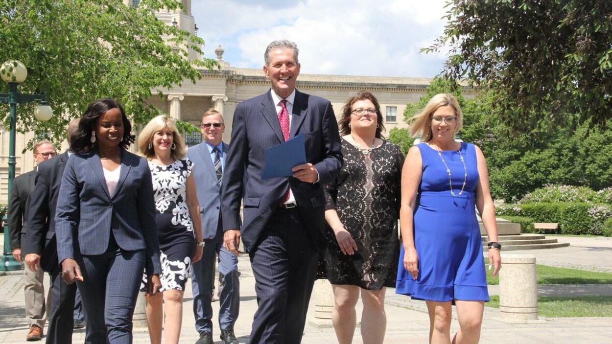 Un très grand homme, Brian Pallister, marche en souriant entouré de femmes, derrière le palais législatif du Manitoba.