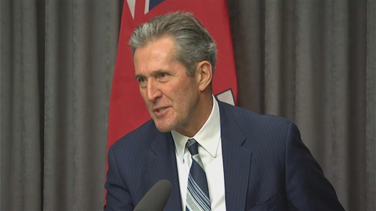 Brian Pallister devant un microphone. Derrière lui, il y a un drapeau du Manitoba.