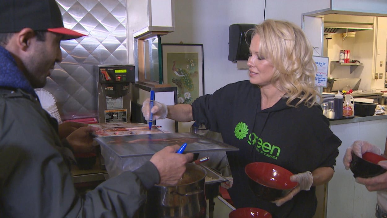 Pamela Anderson signe une affiche, un bol de nourriture dans les mains. Elle se trouve dans une soupe populaire.