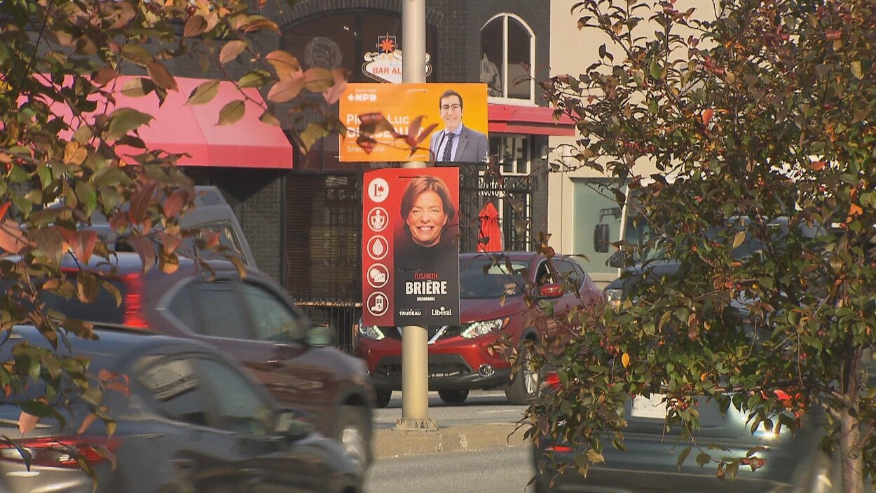 Deux pancartes sont superposées sur un poteau installé sur un terre-plein d'un boulevard où passent des voitures. Sur une des pancartes, on retrouve la photo d'un homme et sur l'autre, la photo d'une femme.