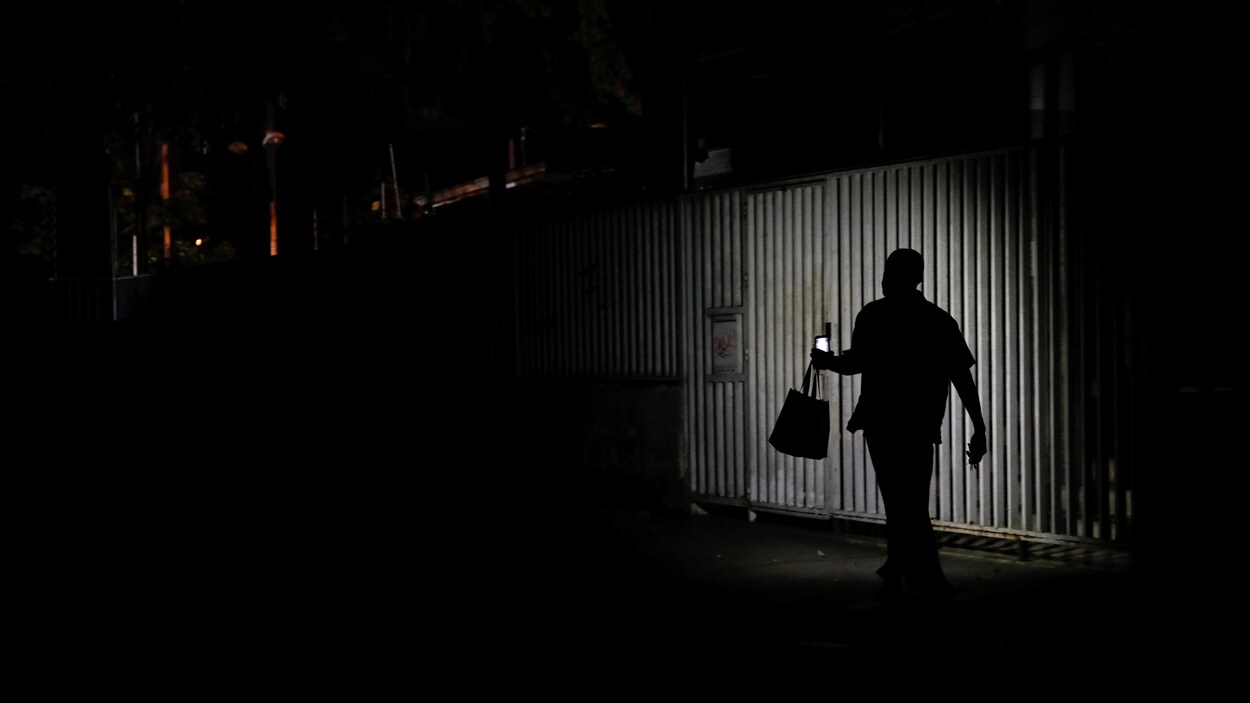 Un individu marche, guidé par la lumière de son téléphone portable, devant une clôture métallique.
