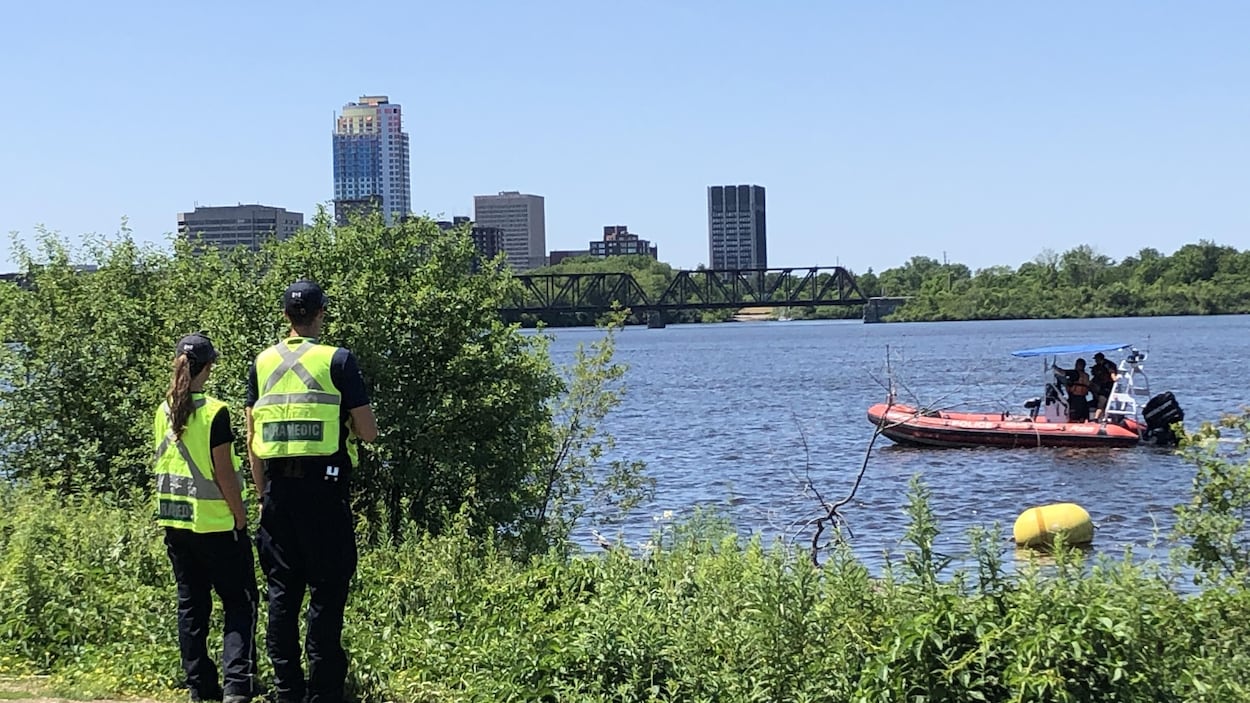 Deux ambulanciers scrutent la rivière des Outaouais sur laquelle circule un bateau des services d'urgence.
