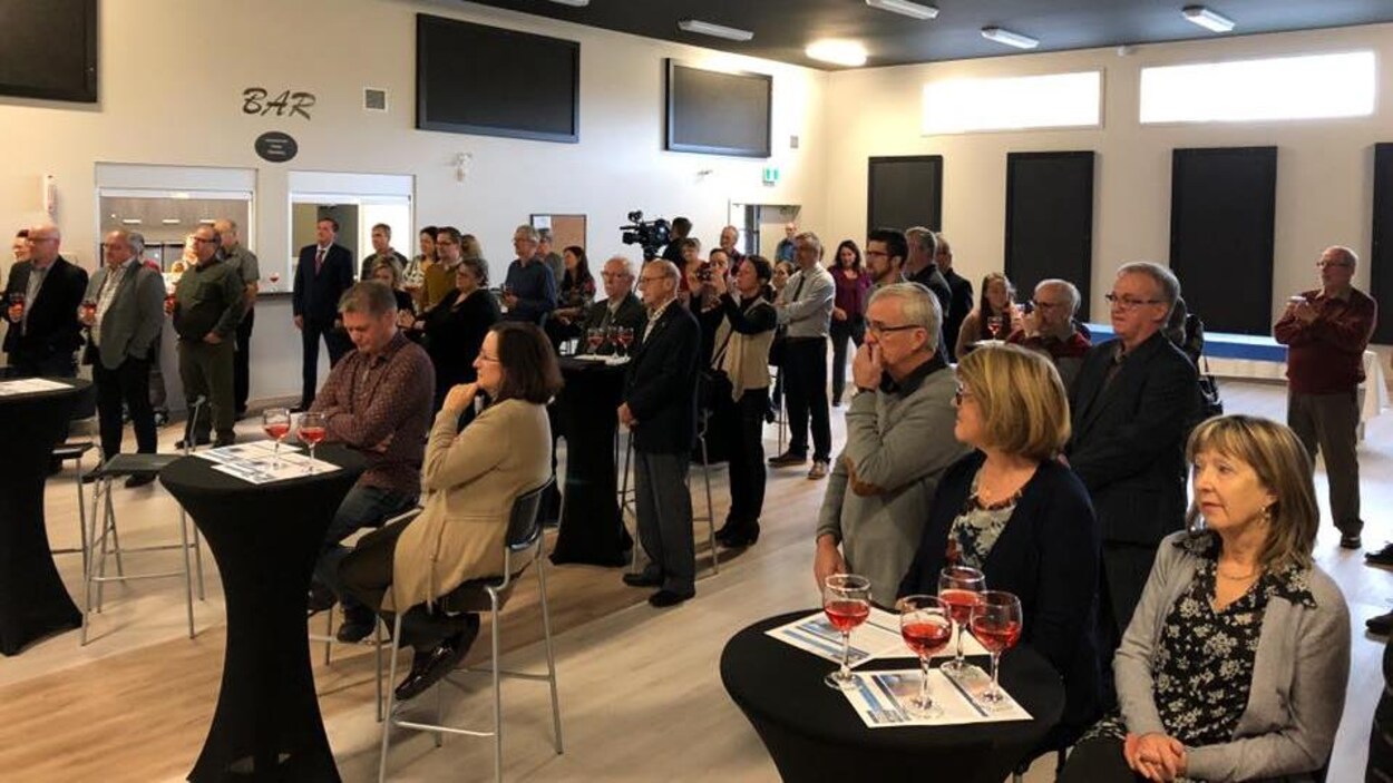 Quelques dizaines de personnes sont rassemblées dans une salle. Des verres de vin sont posés sur de petites tables noires.