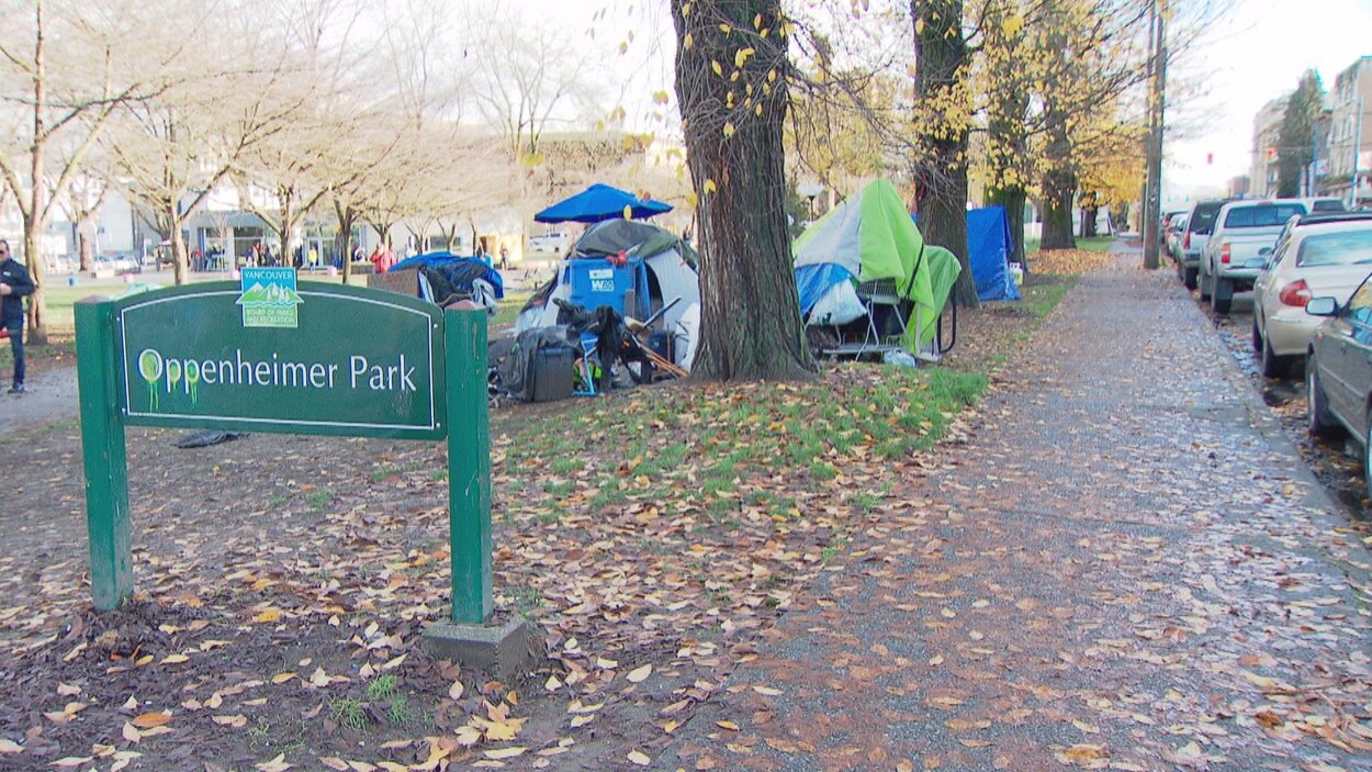 Un panneau inscrit Oppenheimer Park, avec des tentes en arrière-plan, dans un parc, et une rue à droite.