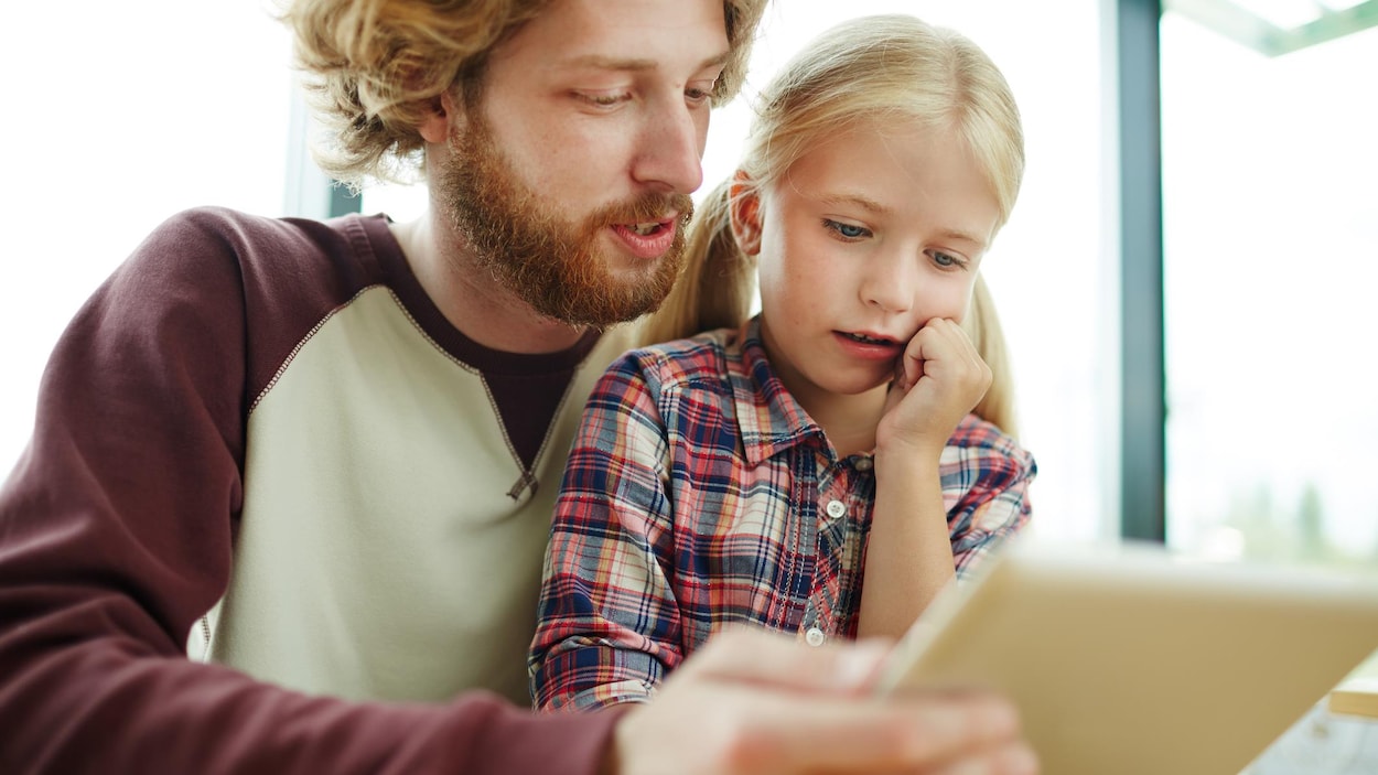 Un père tient une tablette électronique et la montre à sa fille.