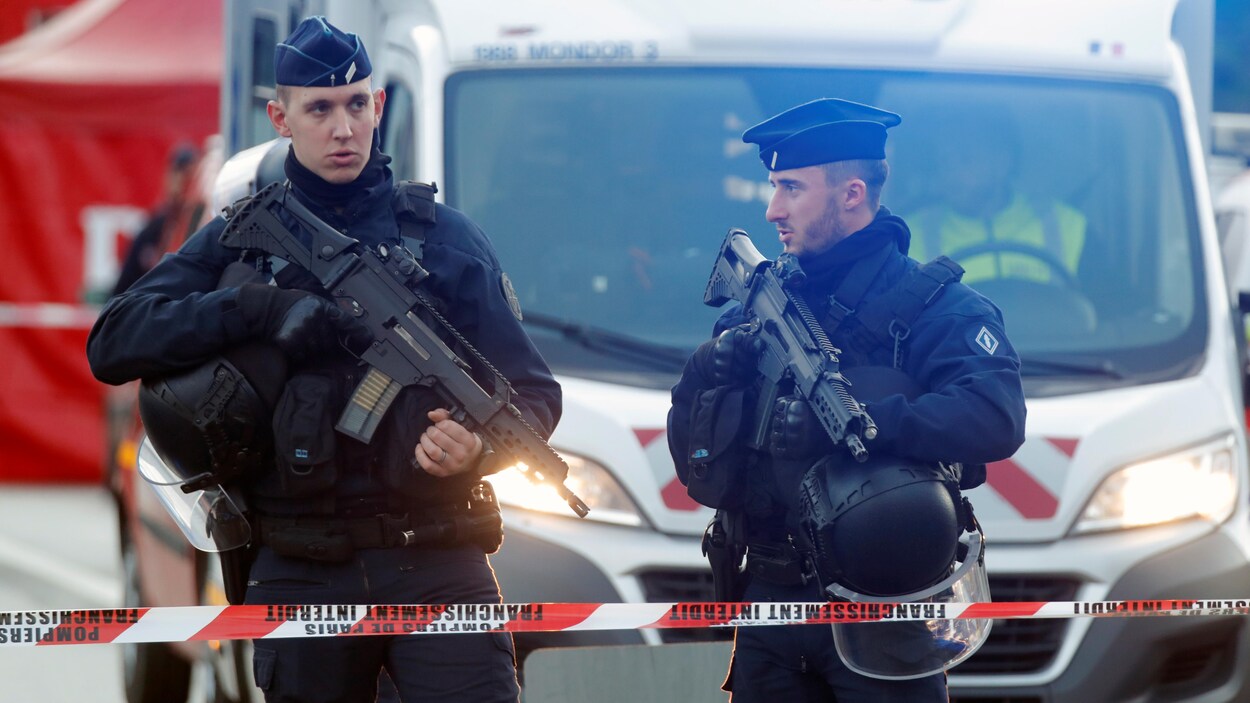 Deux policiers armés derrière un ruban rouge et blanc et devant une ambulance sur laquelle est écrit SAMU.