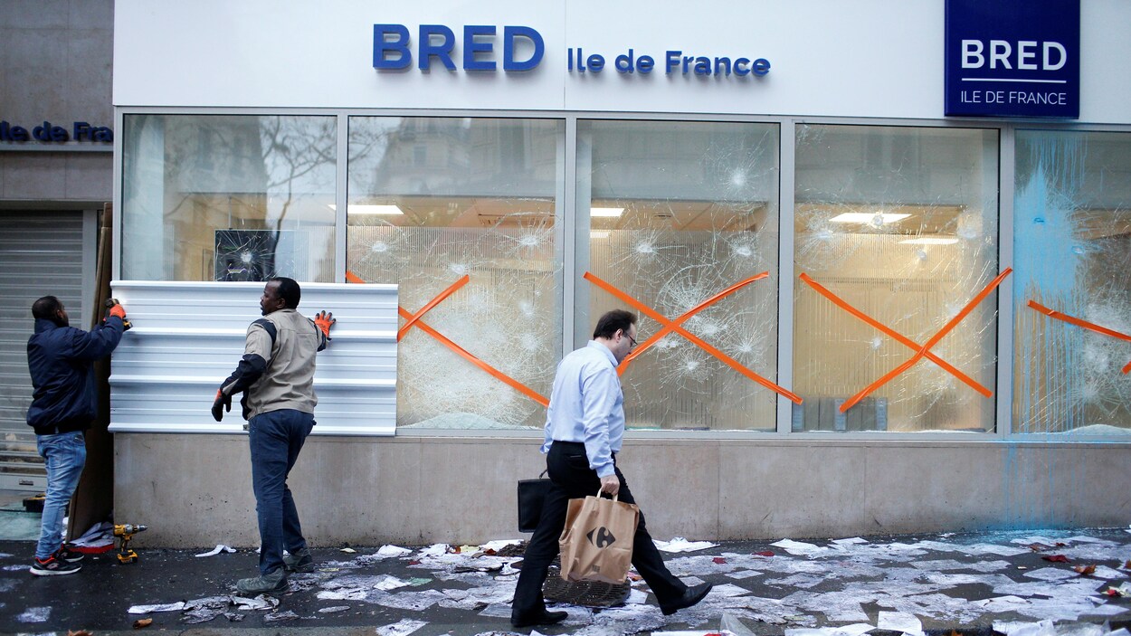 Deux hommes placent des panneaux devant des fenêtres brisées, alors qu'un homme passe sur le trottoir devant la banque, qui est envahi de déchets.