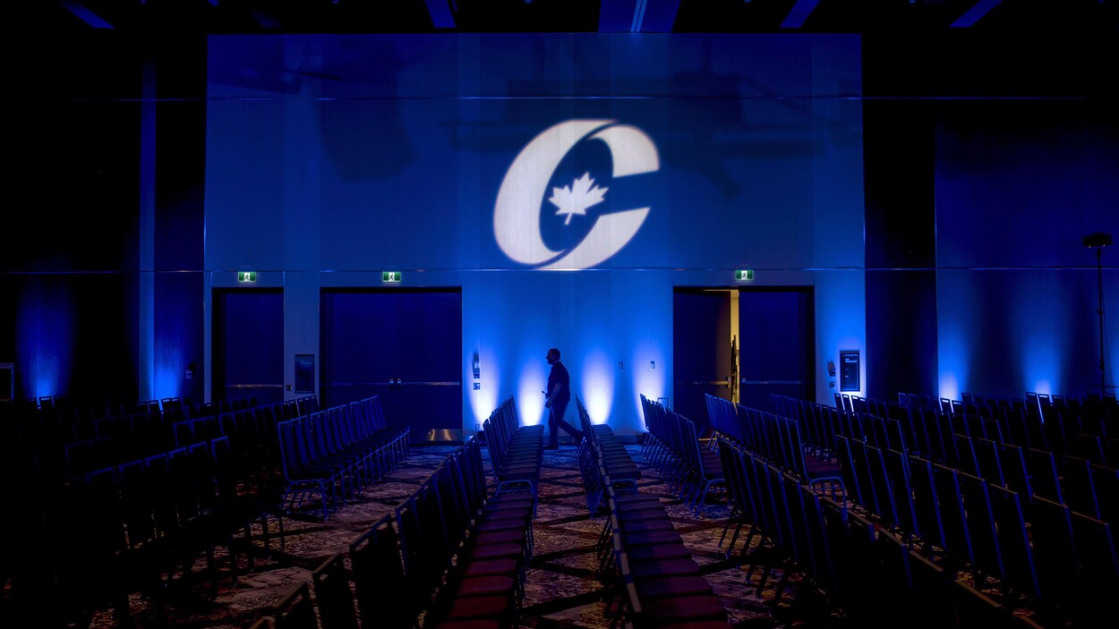 Une salle remplie de chaises vides. Le logo du Parti conservateur du Canada est projeté sur le mur.