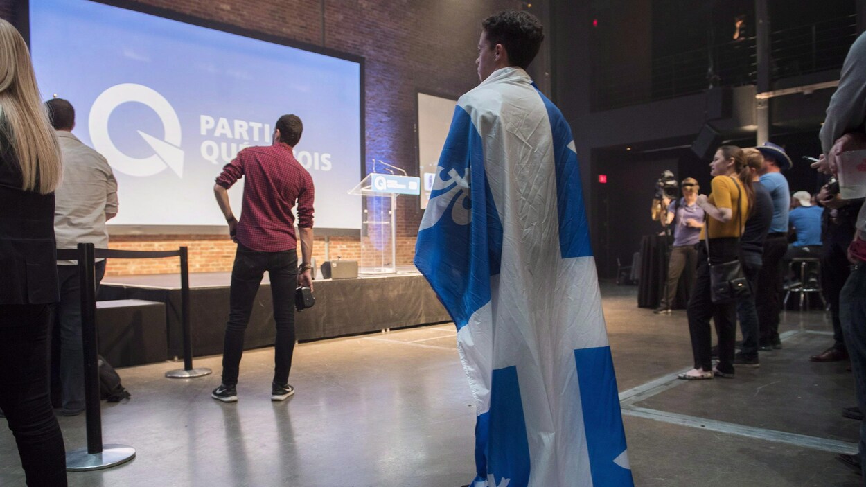 Un militant péquiste, portant le drapeau québécois comme une cape, est debout devant une estrade vide.