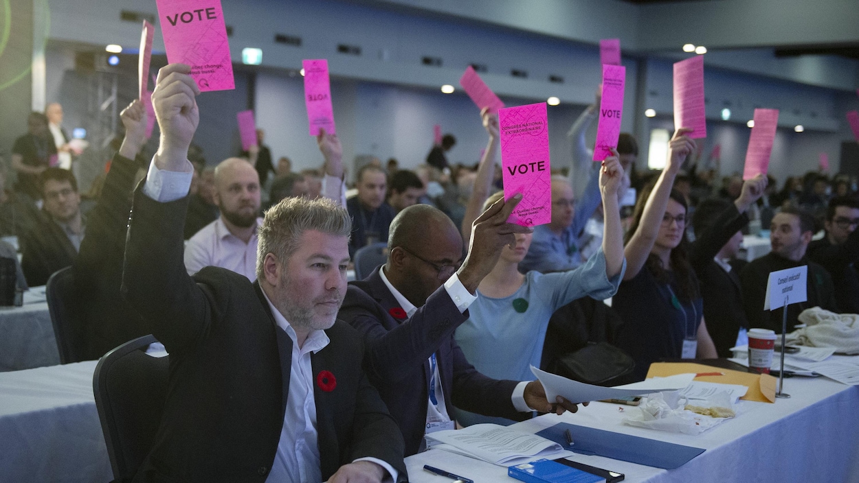 Les membres du parti sont assis dans une salle. Certains tiennent une affiche rose dans les airs pour indiquer leur appui à une proposition.