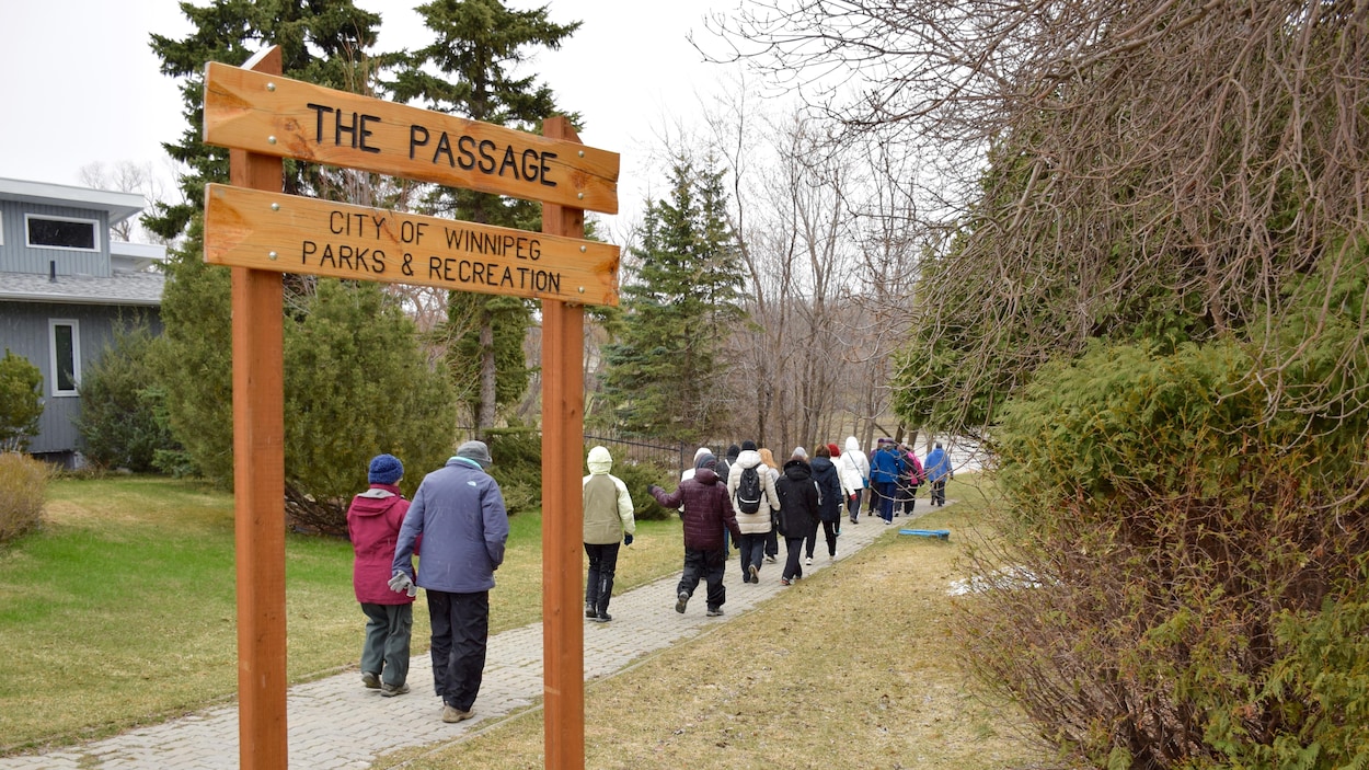 Un groupe de marche suit un trottoir vers la rivière d'où une affiche de la Ville de Winnipeg indique « The Passage ». Un groupe de marche suit un trottoir vers la rivière d'où une affiche de la Ville de Winnipeg indique « The Passage ».
