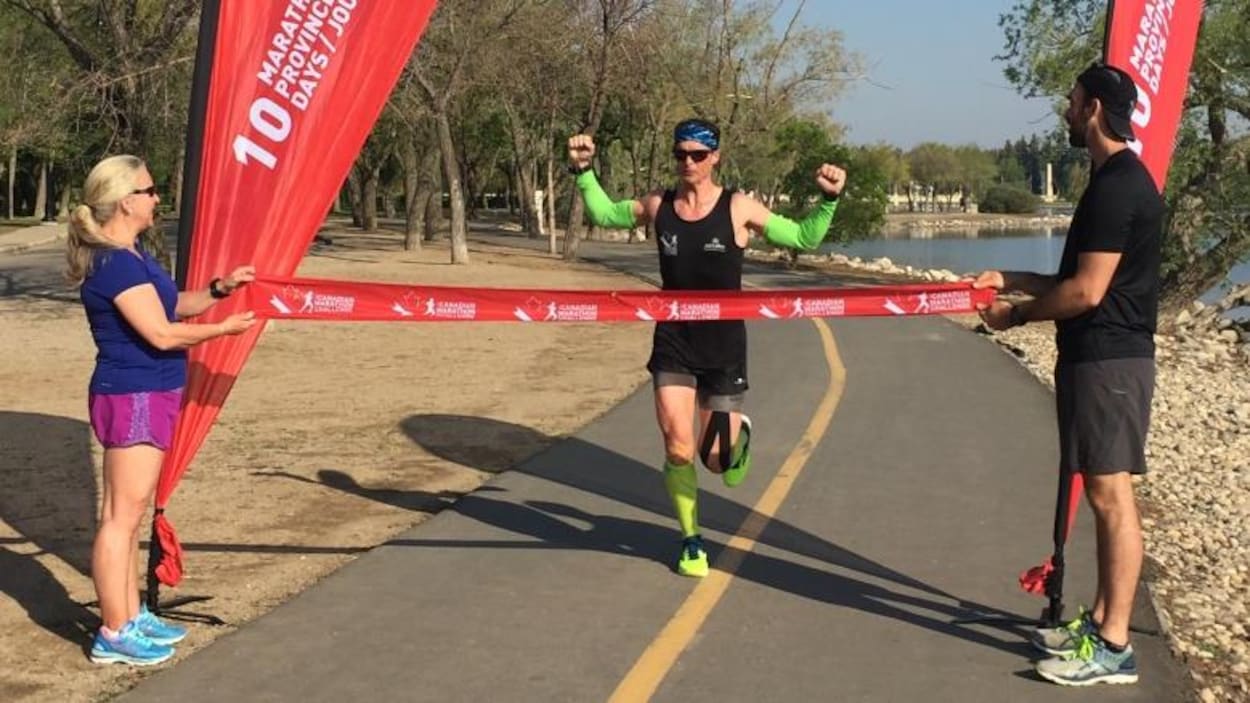 Patrick Charlebois termine son marathon autour du lac Wascana à Regina en levant les bras à la ligne d'arrivée sous un ciel bleu.