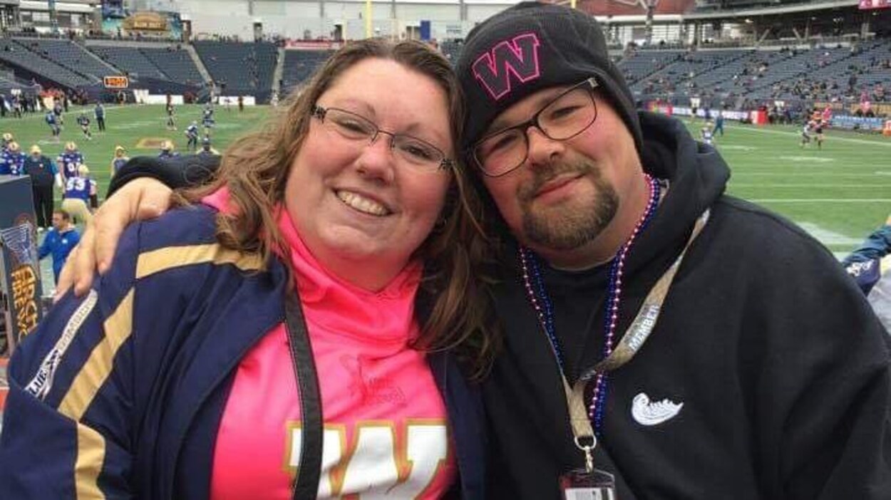 Une femme et un homme, Danielle Vandale et Patrick Gagnon. sourient à la caméra. Ils sont dans le stade de football des Blue Bombers à Winnipeg et portent des vêtements aux couleurs de leur équipe.