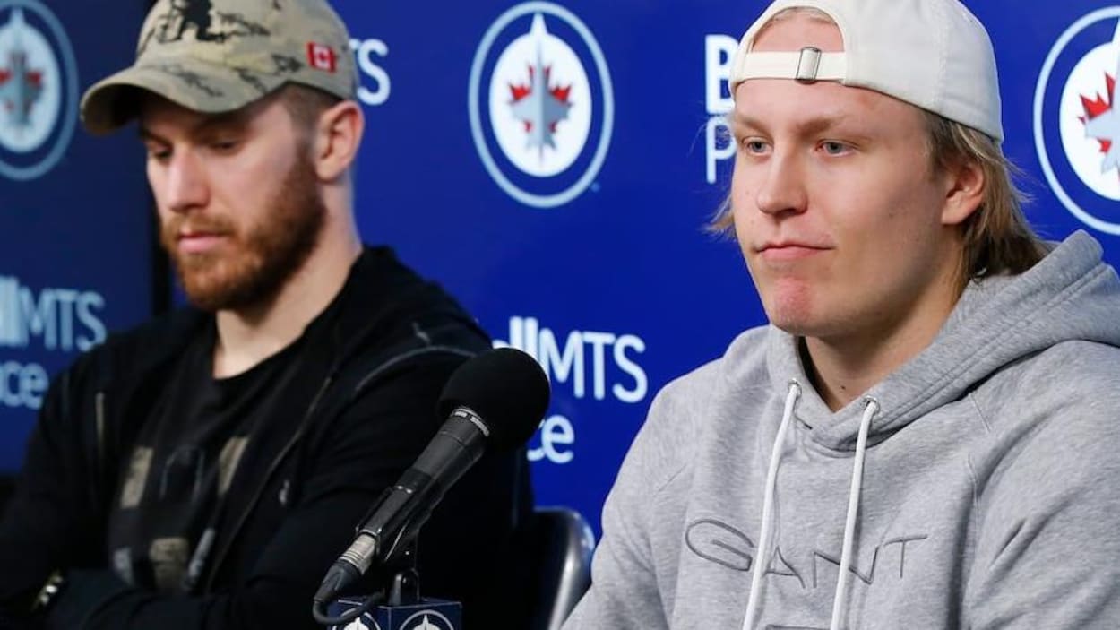 Deux joueurs de hockey sont assis devant des micros et parlent aux journalistes. L'un d'eux est Patrick Laine, qui est vêtu d'un chandail de coton ouaté et d'une casquette.
