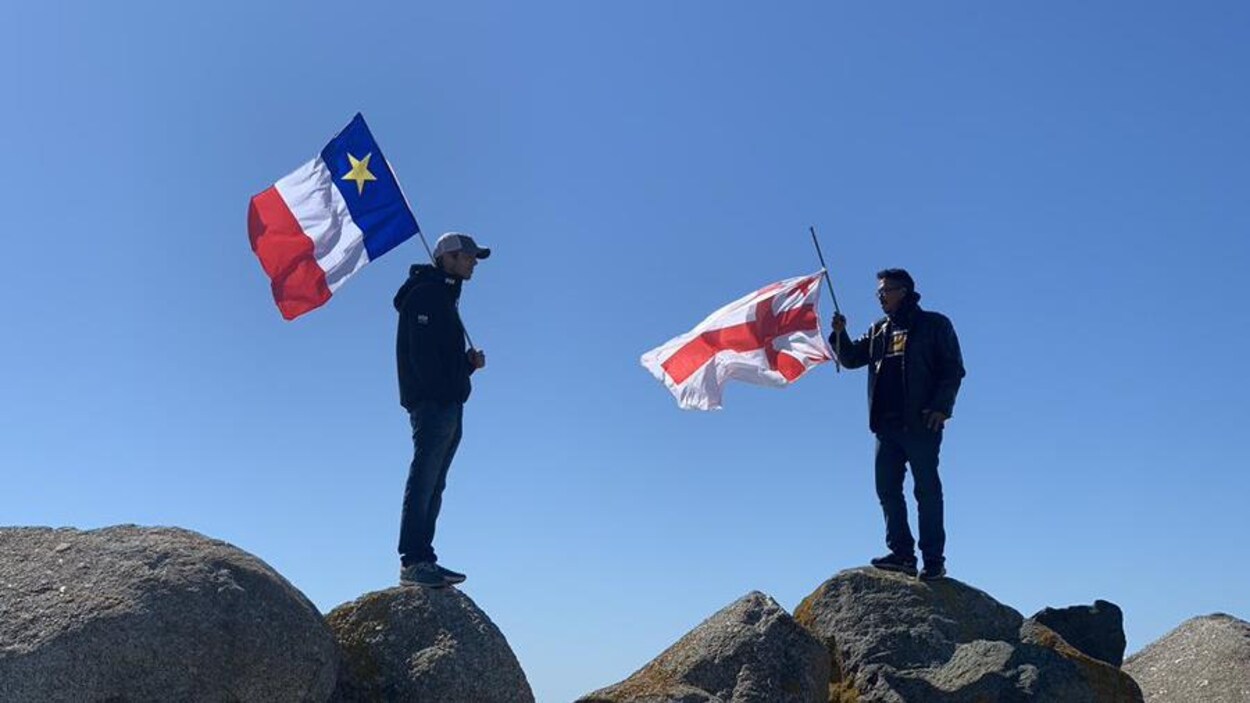 Deux hommes debout, chacun sur un rocher. Ils se font face. L'un a un drapeau acadien et l'autre, un drapeau mi'kmaq.
