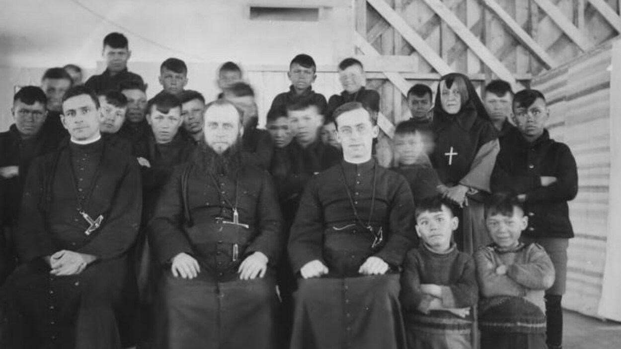 Plusieurs dizaines d'enfants autochtones posent debout. À l'avant de la photo, trois prètres sont assis.