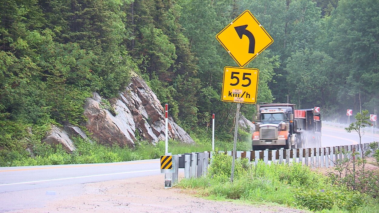 Un camion lourd circule sur une route. À l'avant-plan, on voit un panneau de vitesse indiquant 55 kilomètres à l'heure.