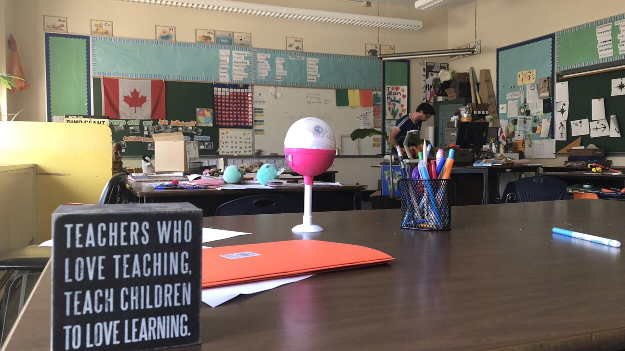 Une table dans une classe avec des objets dessus et un enseignant à son bureau.
