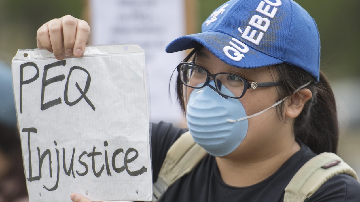 Une femme portant un masque sanitaire et une feuille sur laquelle on peut lire «PEQ injustice».