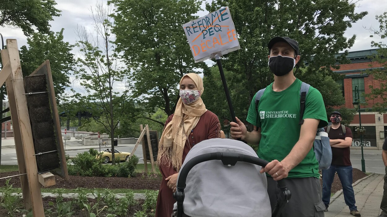 Deux personnes masquées portent une pancarte indiquant « Réforme du PEQ, décalée ».