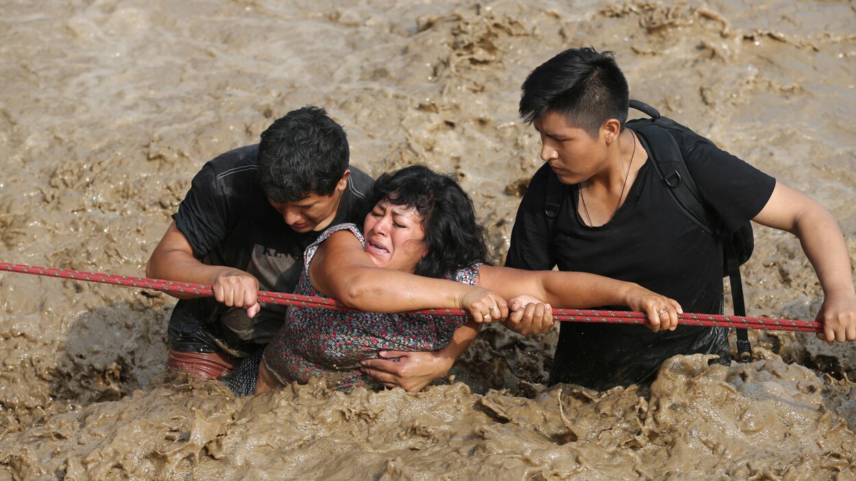 Deux personnes aident une femme à traverser une rue inondée après le débordement d'une rivière à Huachipa, au Pérou. Deux personnes aident une femme à traverser une rue inondée après le débordement d'une rivière à Huachipa, au Pérou.