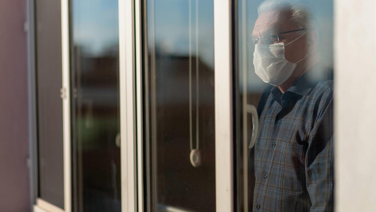 Un homme âgé portant un masque regarde à travers une fenêtre.