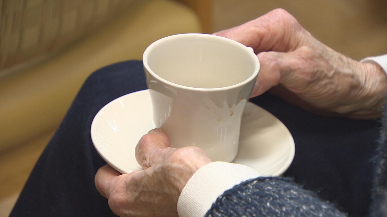 Une personne âgée, dont on ne voit que les mains, boit un café d'une tasse en procelaine.