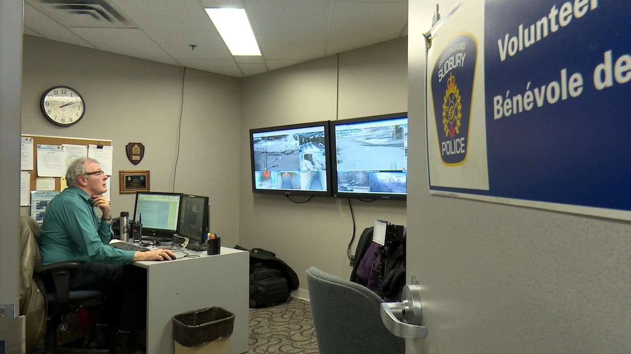 Peter Marshall, assis à un bureau avec en face de lui plusieurs écrans sur lesquels on voit des images du centre-ville, captées par des caméras de surveillance.