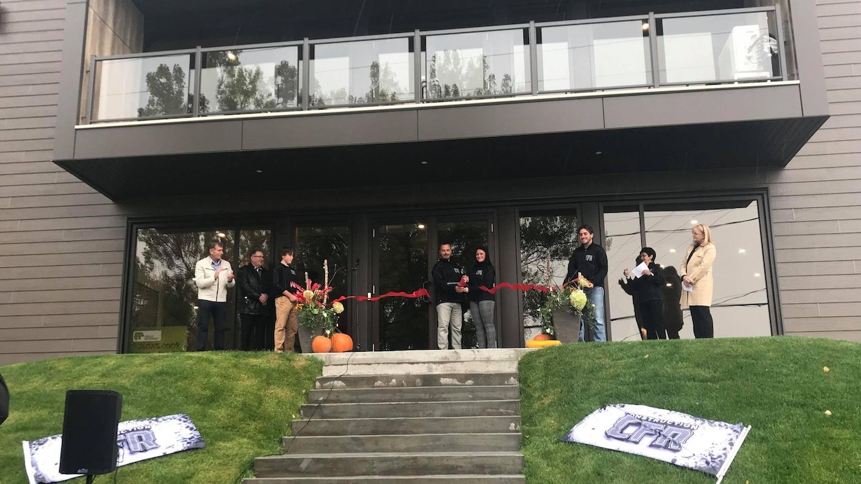 Un homme et une femme coupent ensemble le ruban. Ils sont accompagnées de d'autres personnes devant l'entrée principale de la résidence.