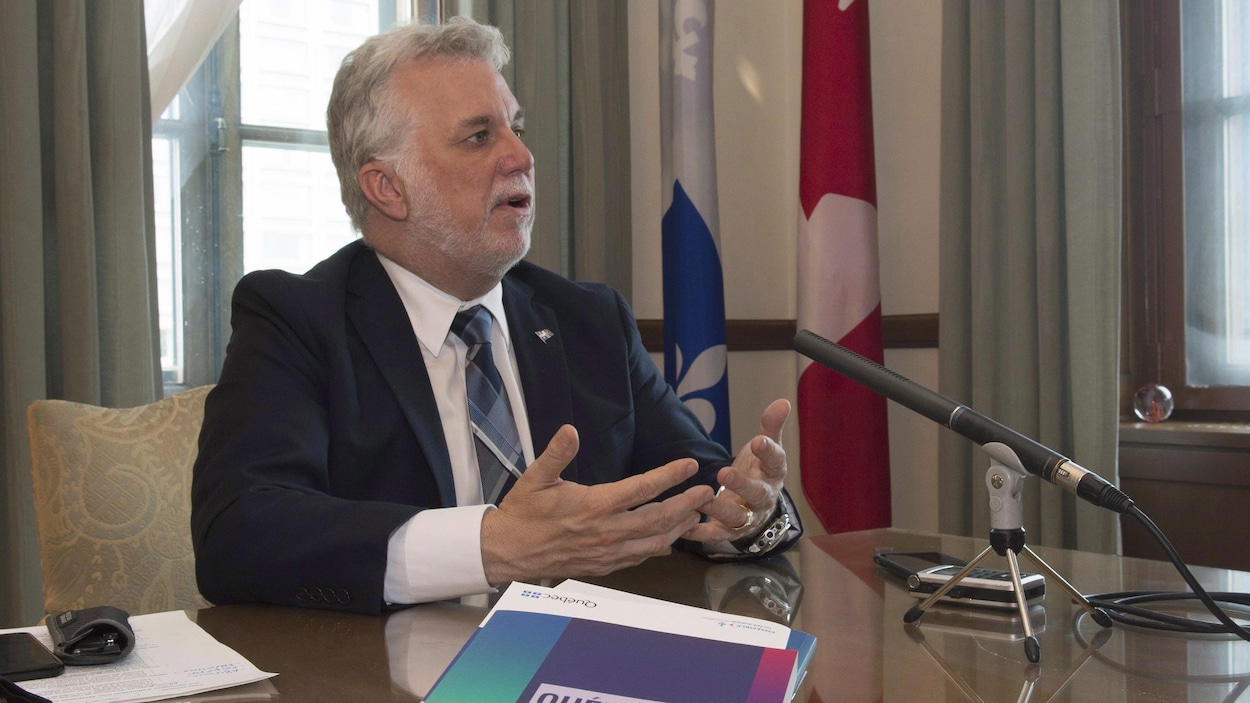 Philippe Couillard, assis derrière un micro, parle aux journalistes. Un exemplaire du document sur la Constitution est posé sur la table devant lui. Philippe Couillard, assis derrière un micro, parle aux journalistes. Un exemplaire du document sur la Constitution est posé sur la table devant lui.
